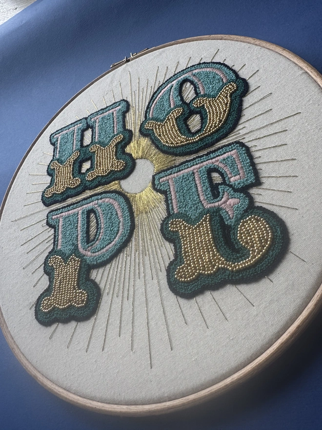 A close up of an embroidery hoop containing the stitched word Hope sewn in shades of green and gold beads with gold floss rays 