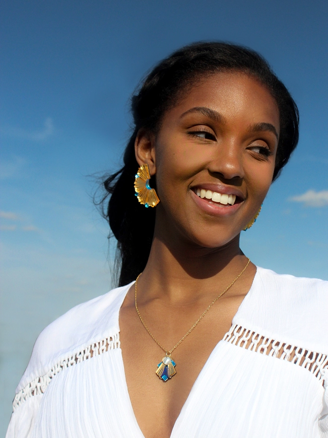 A portrait of a smiling model wearing an earring that resembles like the coral wall from Lamu Island and a blue necklace. Her hair is flowing behind her. 