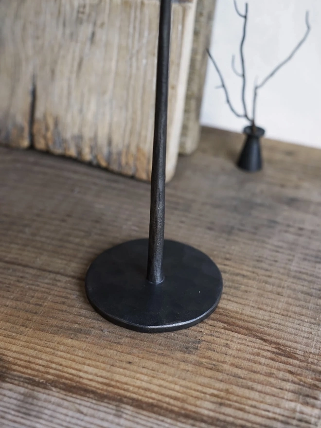 Forged Steel Candle Stick, a metal candle stick sitting on a wooden surface against a wood backdrop. 