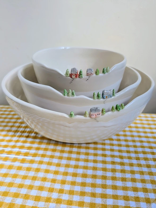 Handmade ceramic white nesting bowls with tiny colourful house and tree details on the rim of all three bowls nesting inside each other  on a yellow gingham table cloth