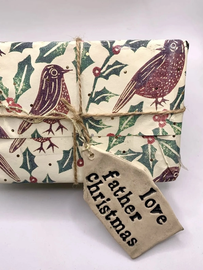 'Love Father Christmas' Handmade Ceramic Gift Tags, a ceramic gift tag attached to a gift sitting on a pale purple surface. 