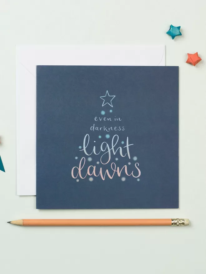 A square dark blue card with a white envelope tucked beneath it lies on a pale blue background. The card features a lettered design which reads: 'Even in darkness, light dawns' in the shape of a Christmas tree