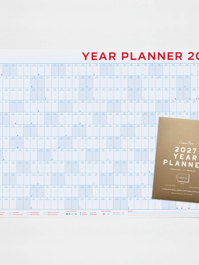A large 2027 wall planner by Crispin Finn, viewed horizontally. The single-sheet calendar features a grid layout for all 12 months, printed on 100% recycled paper using a striking retro two-tone red and blue color scheme, with weekends prominently highlighted in red, with the brown kraft screen printed packaging laid on top