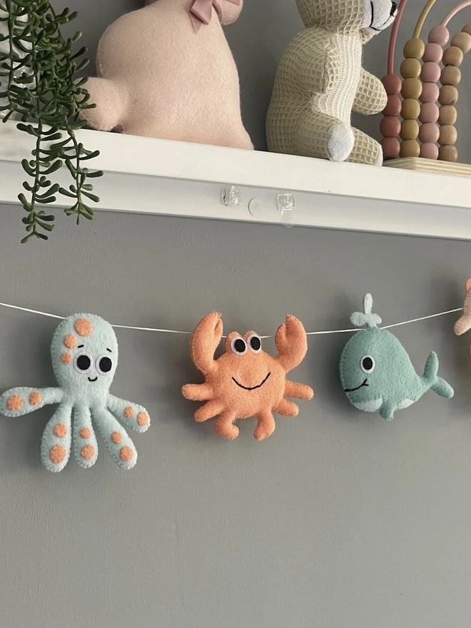 Nursery Under The Sea Garland, a sea themed garland hanging from a white shelf against a grey backdrop in a child's bedroom. 