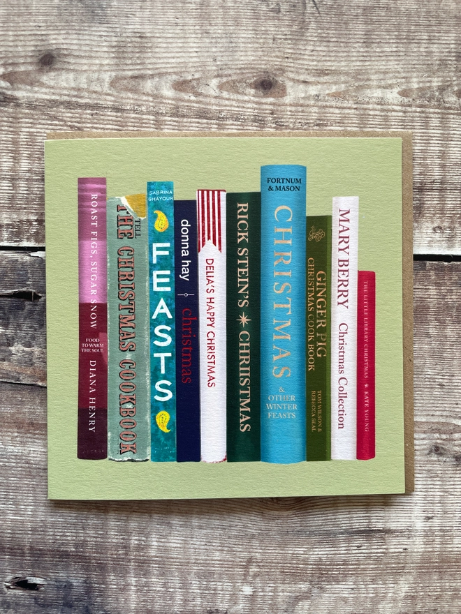 Green Christmas card illustrated with a stack of colourful cookbook spines, including titles styled after Nigella's Feasts, Delia's Happy Christmas, Rick Stein's Christmas, Mary Berry's Christmas Collection, and Fortnum & Mason Christmas, on a rustic wood background.