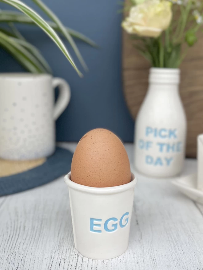 A handmade ceramic egg cup, emulating a paper cup, has the word ‘egg’ painted in blue on its side.