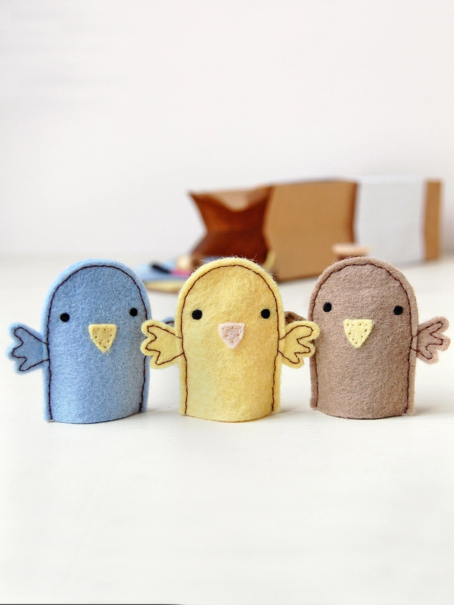 Three felt bird finger puppets stand on a white desk in from of a craft kit box. There is one yellow, on brown, and one blue bird.