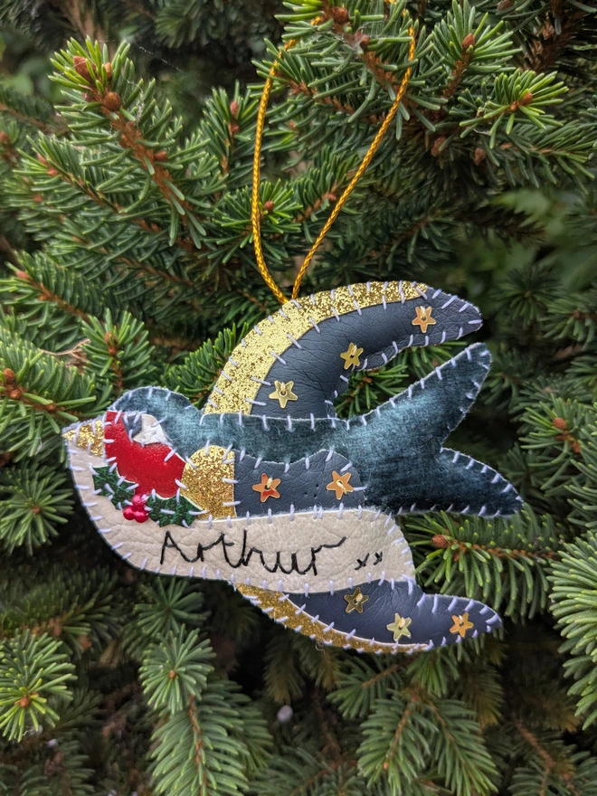 Hand stitched personalised swallow Christmas decoration made from vegan-friendly faux leather and velvet, featuring gold stars, festive holly and a cream banner personalised with a name, hanging from a gold cord on a Christmas tree.