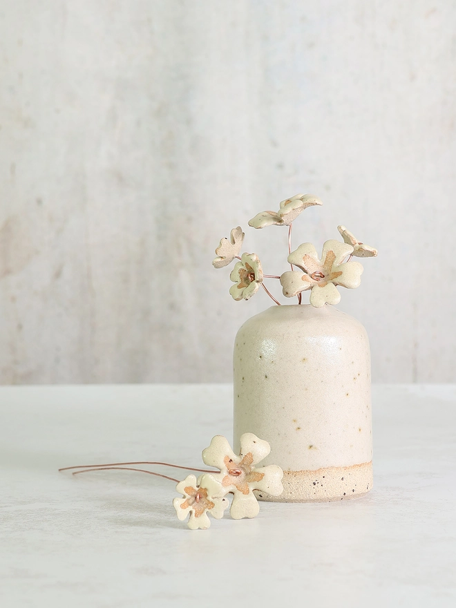 a white table top with an off white handmade stoneware vase sitting on top, the vase displays a small posy of ceramic primroses in a soft yellow colour. Beside the vase sits lays two flower stems 