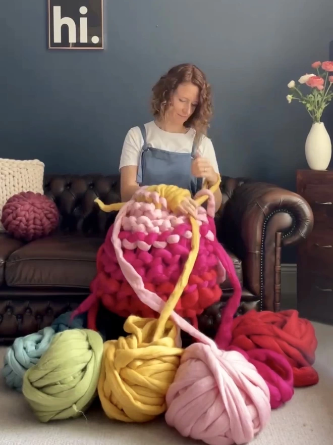 Mizz, a white woman in her forties, is sitting on a brown leather sofa. She is looking down at her arms as they knit a large rainbow coloured merino blanket, the balls of giant yarn is bright colours sit in the foreground