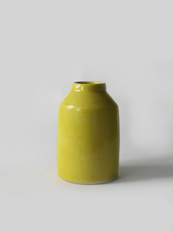 A small handmade ceramic bud vase in glossy sun yellow glaze, with a rounded bottle shape and narrow neck, photographed against a plain white background