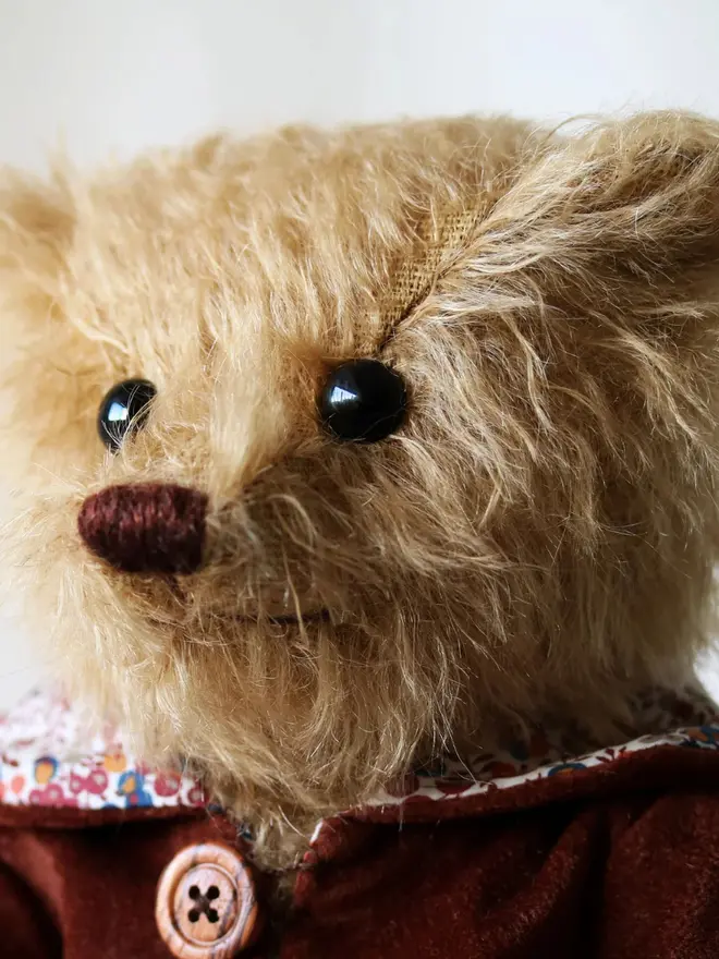 Brown teddy bear wearing a burgundy cloak with flower details and a brown button, sitting upright against a white background.