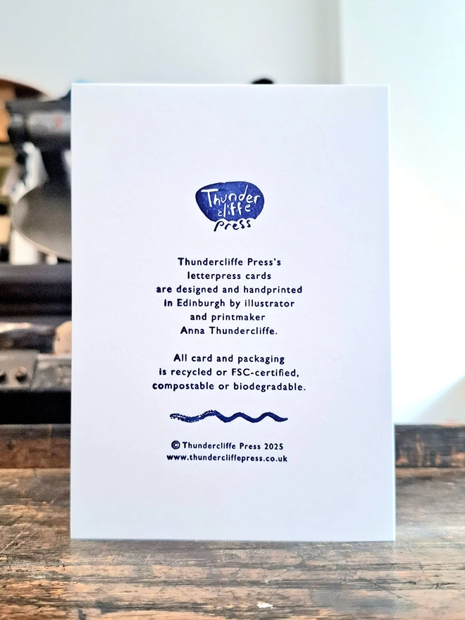 The back of a greetings card showing blue handprinted text, standing on a wooden surface.