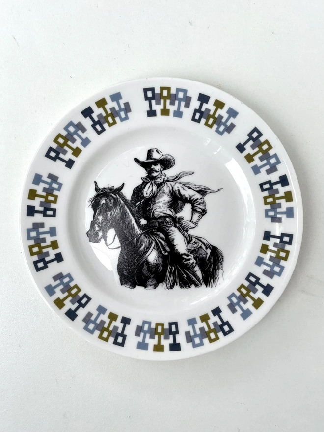 vintage plate hanging on a white wall, it has a decorative border of grey and blue leaves, with a printed vintage illustration of a cowboy in the middle 