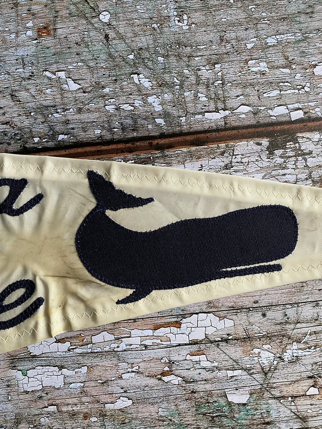 Detail of an up cycled sail cloth 'Keep the sea plastic free' pennant flag showing the detail of the navy canvas whale on the end on the pennant.