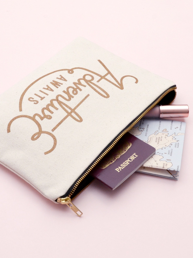 a natural canvas zip pouch with the words adventure awaits on a pink background with a passport and map spilling out 