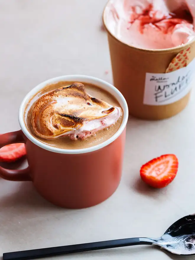 Strawberry Wonder Fluff Tub, looking down on a hot chocolate topped with torched strawberry fluff, in front of an open tub. This is on a marble surface with fresh strawberries scattered around.