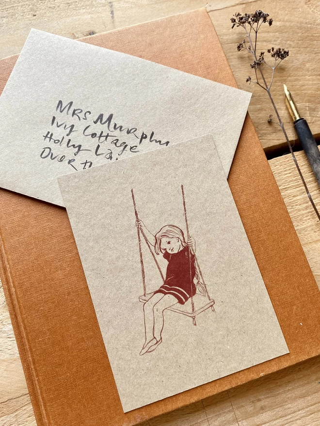 a little girl on a swing printed in terracotta on a kraft postcard shown with a kraft envelope