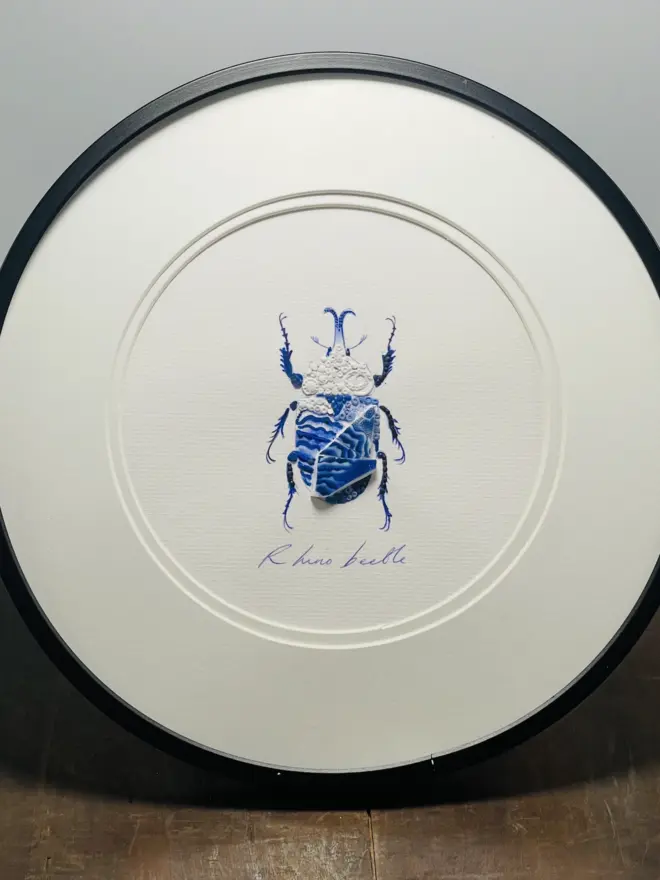 Hand-drawn, painted, mosaic clay, vintage blue Chinese Willow Tree pottery insect on white double mounted card, in a black circular frame.