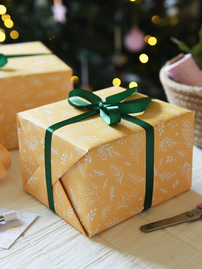 Gifts wrapped in yellow wrapping paper with a gentle botanical design and tied with a green ribbon are on a table in front of a Christmas Tree.