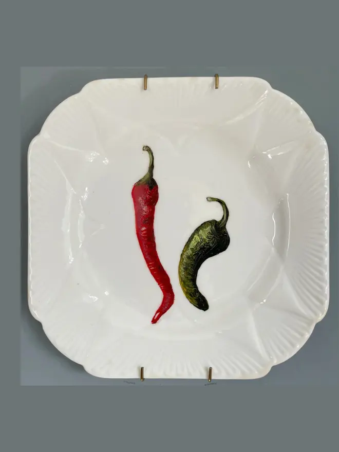Oil Painted Two Chilli On A Tea Plate, hanging on a grey wall.