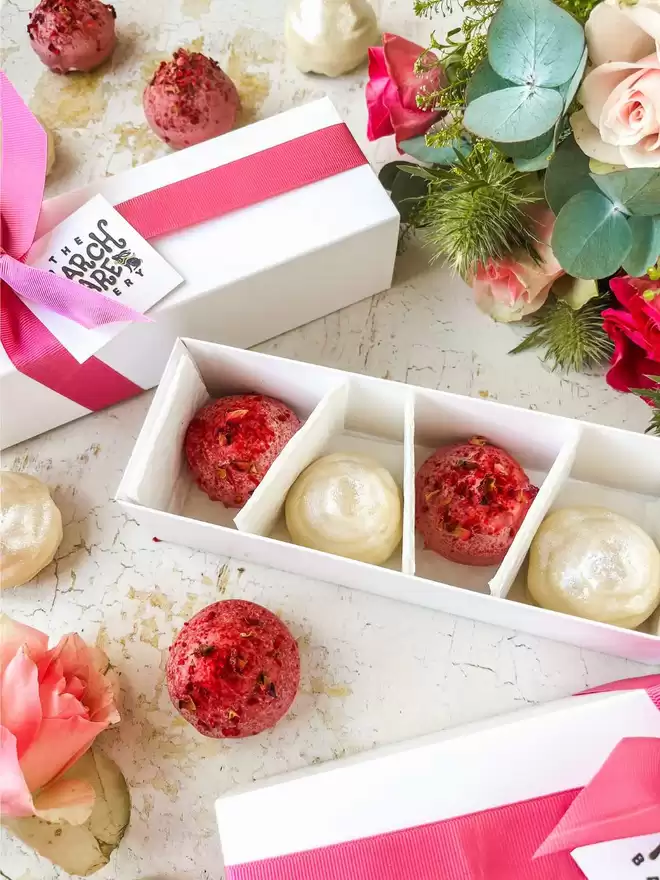 white boxes filled with chocolate covered marshmallow and macaron treats shaped like little kisses and tied with pink ribbons sit on a table next to a bouquet of flowers