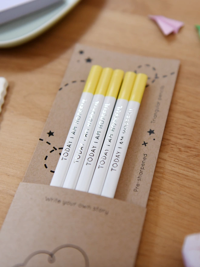 Five white and yellow pencils with the words Today I Am Hopeful along the sides, sit in cardboard packaging on a wooden desk.
