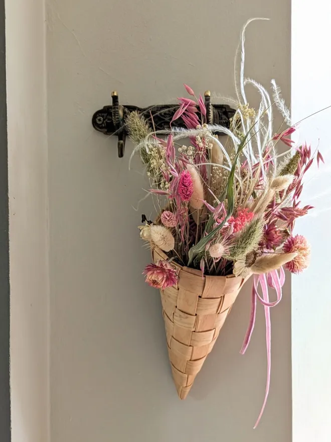 wooden braided cone basket with dried pink flowers hanging from a hook on a wall next to a green coat