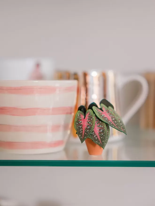 A miniature replica Caladium Red Flash paper plant ornament in a terracotta pot sat on a wooden log slice with a 2nd Caladium sat to the left against a white background