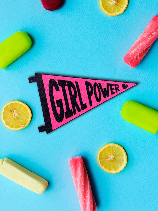 image shows a vibrant blue background with pink and green ice lollies and slices of fresh lemon. in the centre is a small felt pennant with the words 'Girl Power' printed in black ink on hot pink felt.