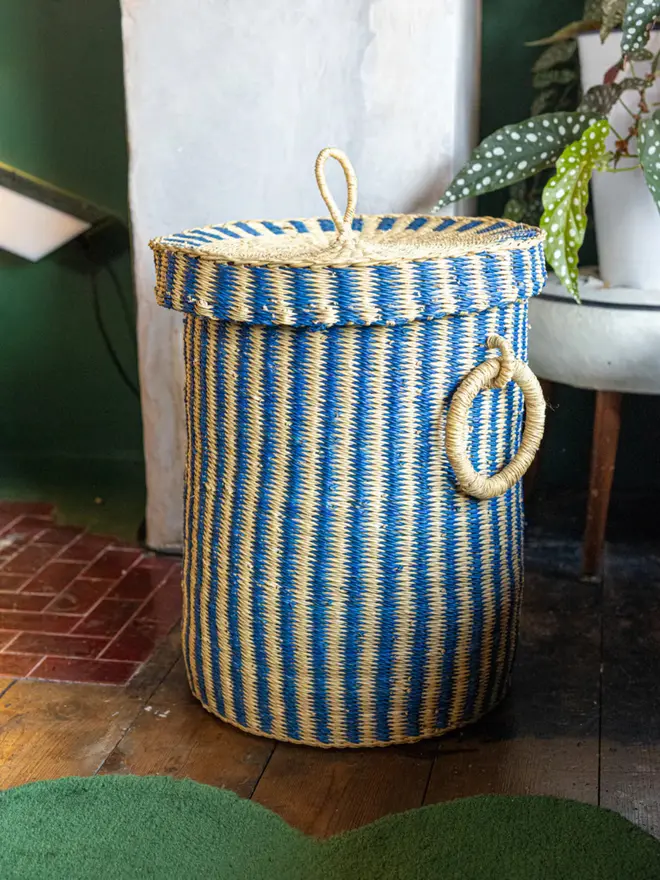 Ghanaian Hand Woven Laundry Basket With Lid And Handles, a colourful basket sitting on a wooden surface in a decorated living room. 