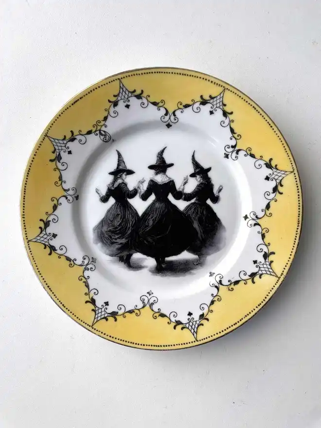 a coven of 3 witches dance hand in hand, the image is victorian in black and white and is printed directly onto an ornate vintage plate