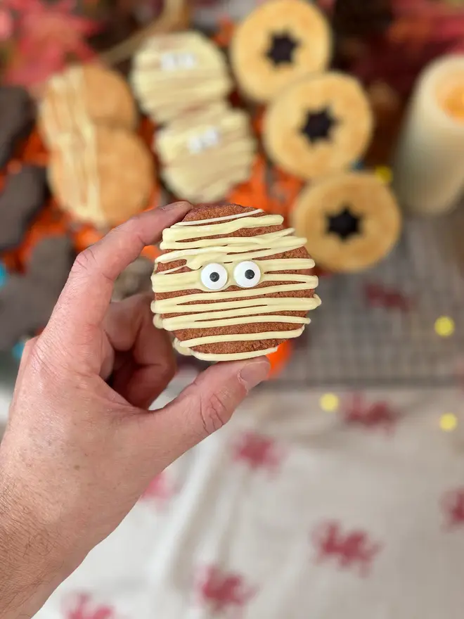A hand holds a mummy-shaped red velvet welshcake decorated with white icing and candy eyes, with other decorated welshcakes blurred in the background.