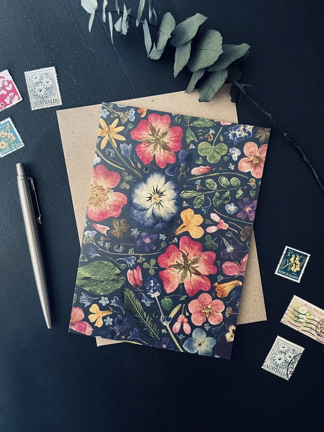 Recycled inky coloured greetings card with digitally printed pressed flower design - Brown envelope - Dark charcoal coloured desk - Colourful flower postage stamps, silver pen, dried eucalyptus