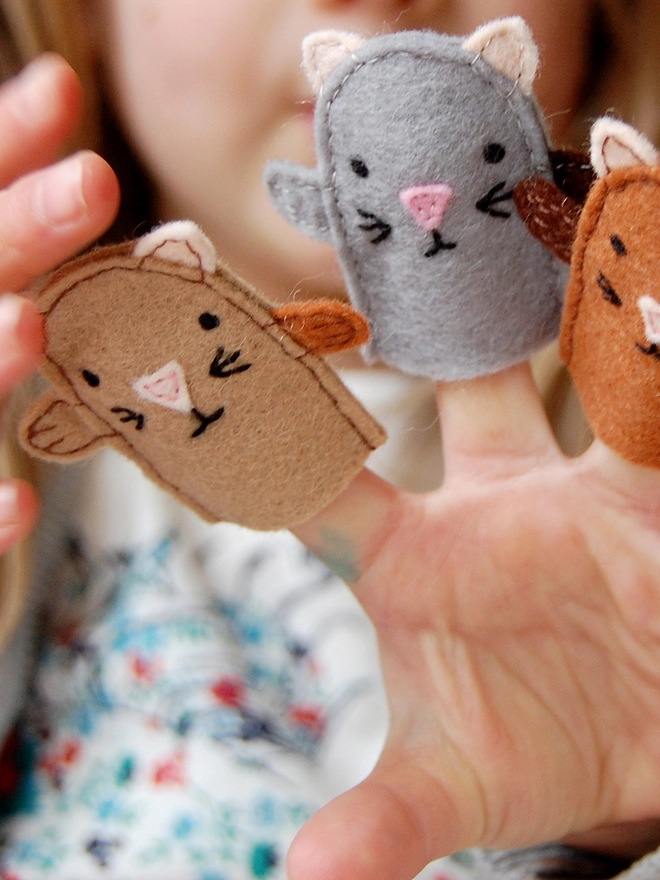 A young child holds three homemade felt kitten finger puppets in her hands.