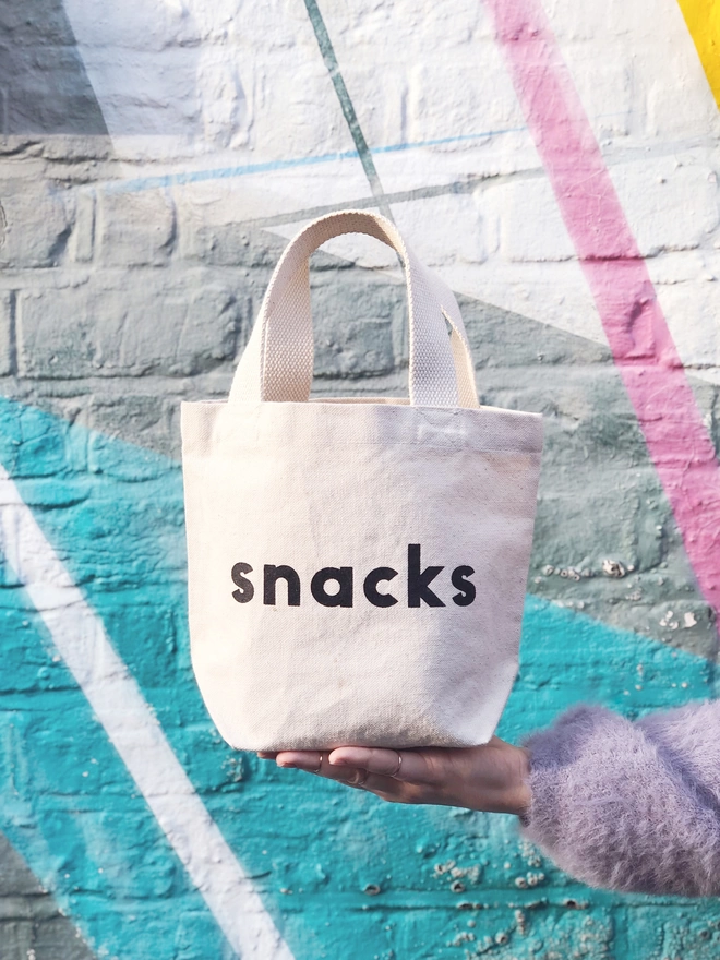 a hand holding out a mini kid's size tote bag with the word snacks on it