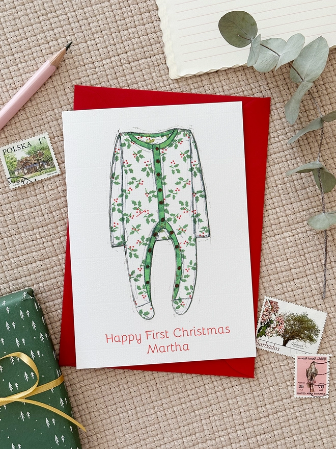 A personalised first Christmas card with an illustrated onesie lays on a red envelope beside various stationery items.