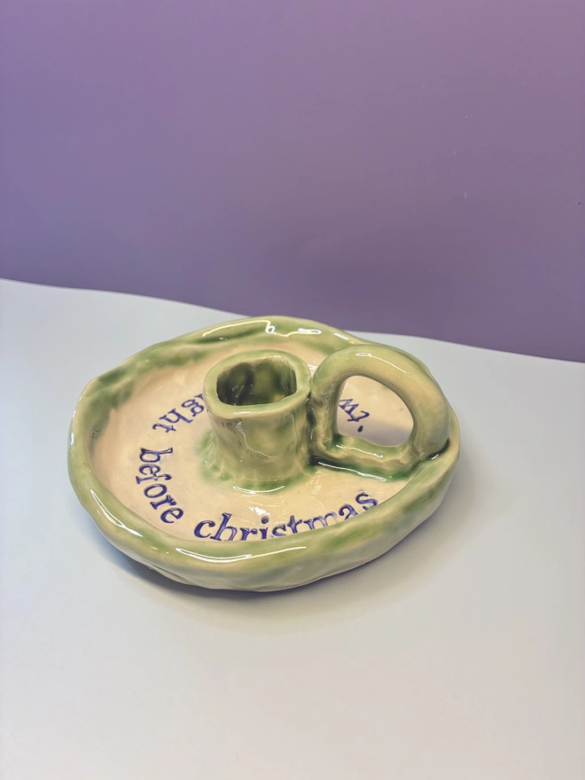 Handmade ceramic Christmas taper candlestick holder with a soft green glazed edge and the words “‘Twas the night before Christmas” in dark blue lettering, arranged around the candle holder opening. Photographed against a pale background.