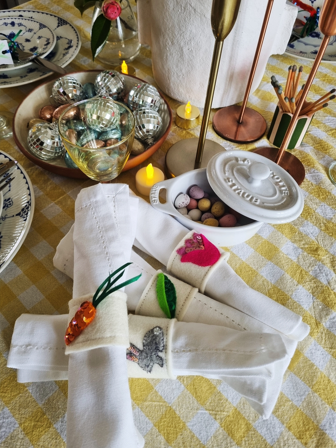 a pile of napkins with the spring set of napkin rings on each one, there are scattered candles, mini eggs in the background