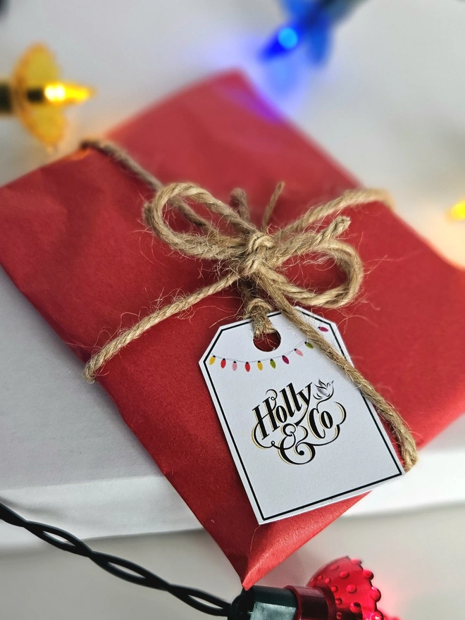 Photograph of the bauble wrapped in red tissue paper with brown twine and a gift tag illustrated with a string of colourful vintage baubles and the Holly and Co logo