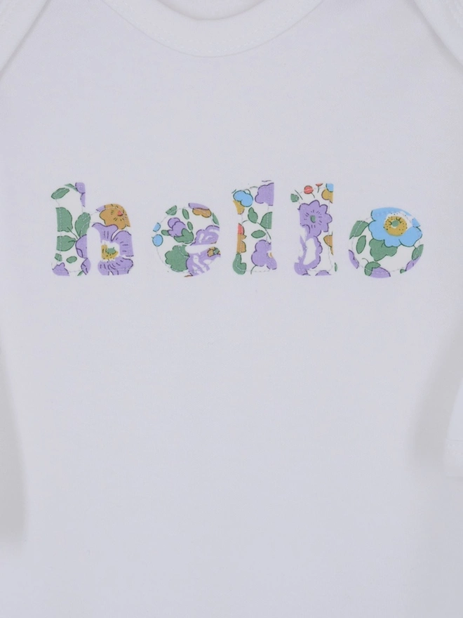 a close up of a white cotton romper with hello sewn onto it in a floral print. close up of the appliqué stitching