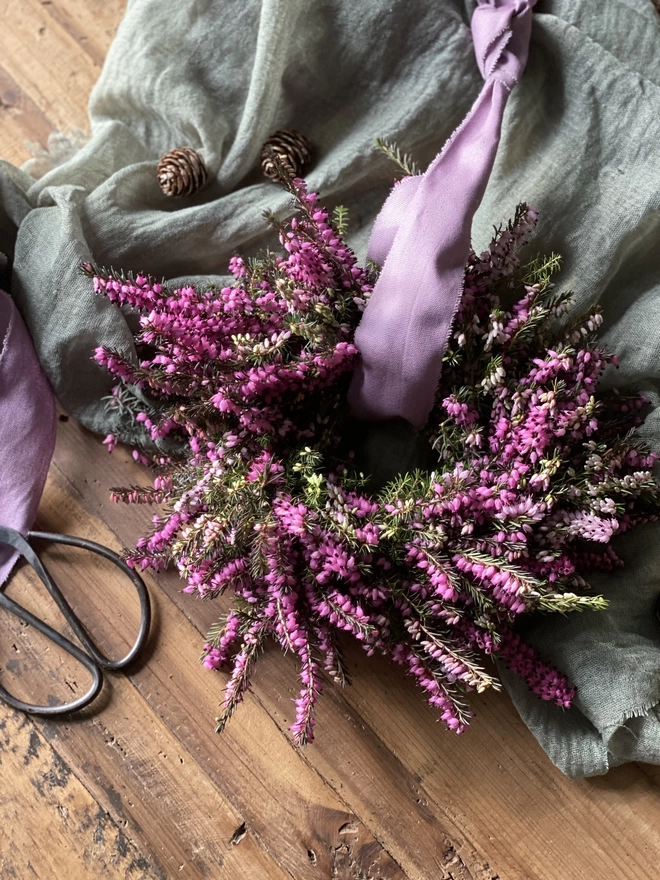 A Petite Wild Heather Wreath in purple and green hues, with a soft lilac ribbon looped through the middle, on display atop a sage green fabric