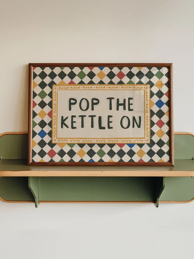 'Pop The Kettle On' checkerboard art print on a wooden shelf on a white and green wall. 