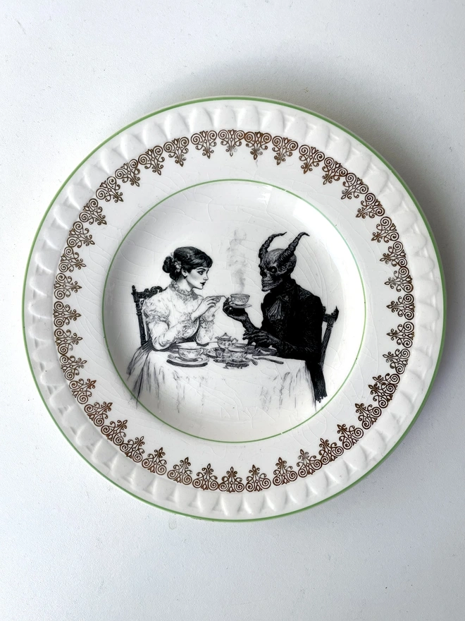 a vintage plate with an ornate edge, ina vintage plate with an ornate edge, in the centre is a vintage illustration of a victorian lady drinking tea with the devil the centre is a vintage illustration of a victorian lady drinking tea with the devil