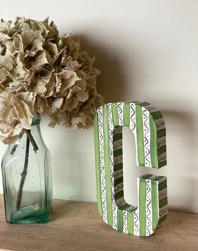 A hand-painted decorative initial letter  with stripes on a wooden table against a light wall. 