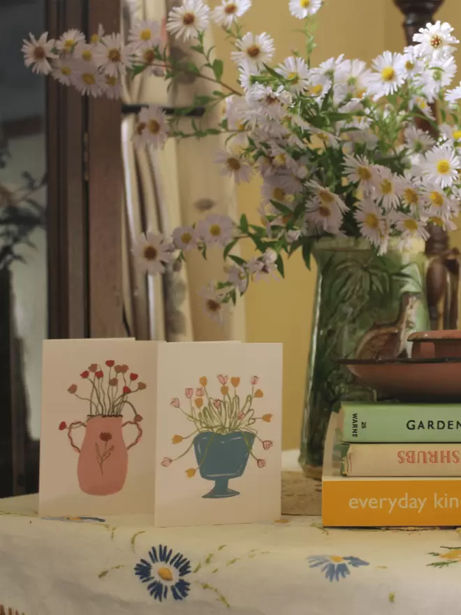 2 cards on a table with flowers on next to gardening books and a bunch of daisies. 