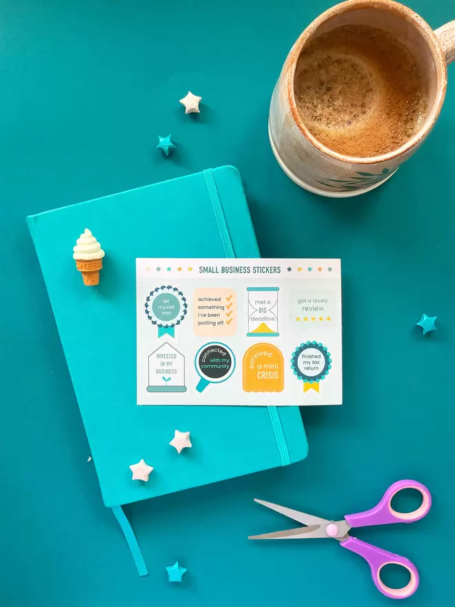 Rock Paper Swan Small Business Achievement Stickers exclusive to Holly & Co seen with a blue notebook, a mug of coffee and scissors.