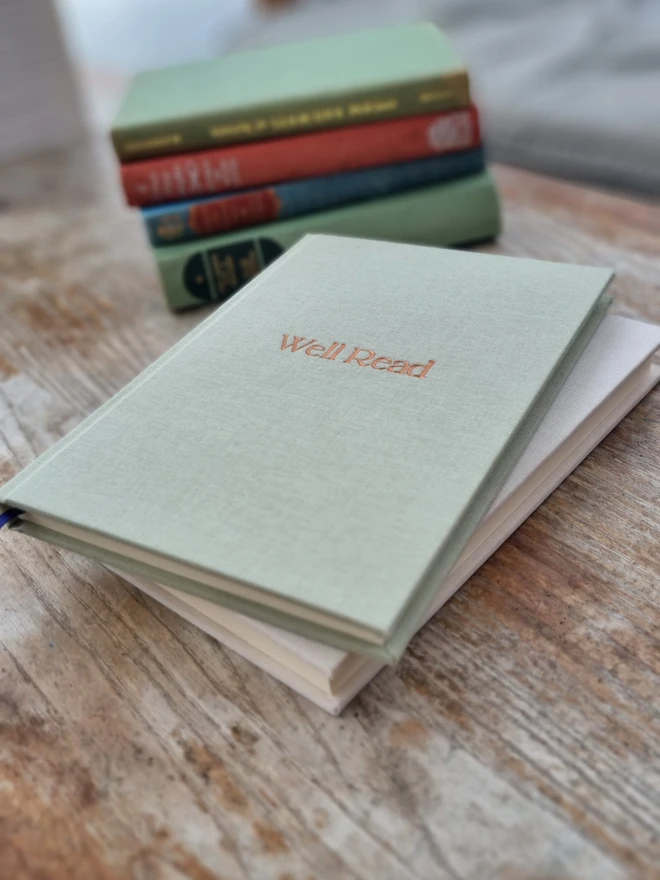 A pale green reading journal with 'Well Read' printed on the cover in gold lettering rests on a wooden table.