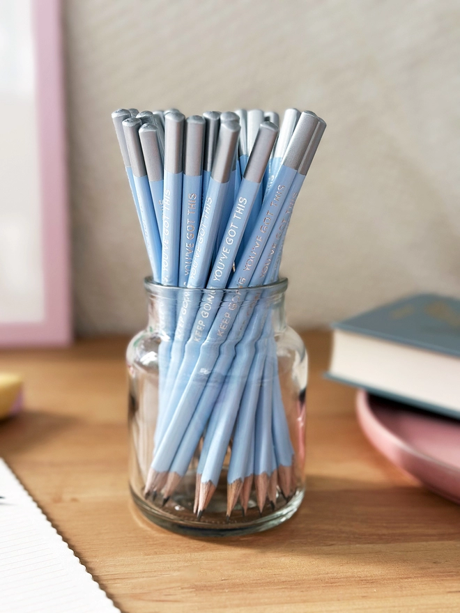 A glass jar full of blue pencils with silver tips stands on a wooden desk. A book lays beside it.