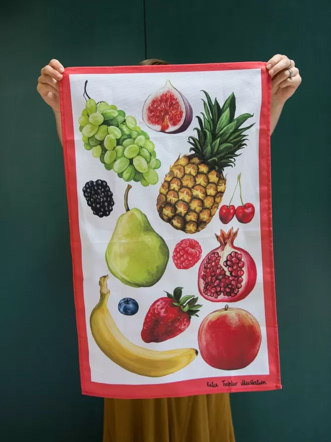 Fruit Themed Tea Towel seen held up in front of a green wall.